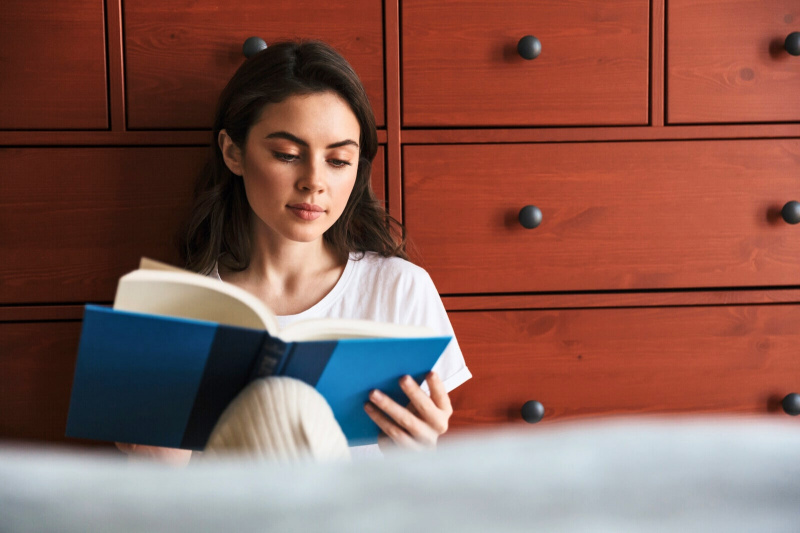 Mujer introvertida sentada en el suelo contra una cómoda leyendo un libro